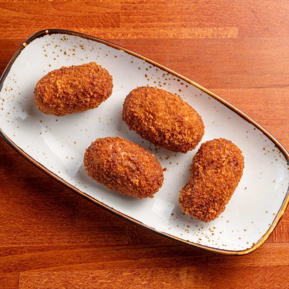 Four golden-brown Spanish croquettes on a white rectangular plate with brown speckles, on a wooden table.