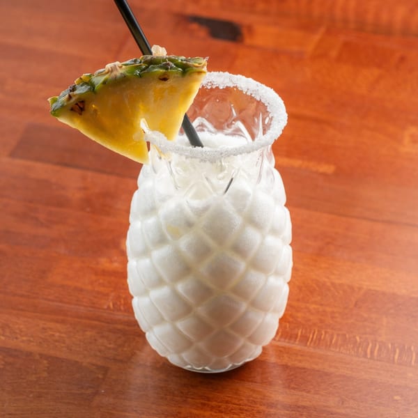 A pineapple-shaped glass filled with a white drink, garnished with a pineapple slice and sugar rim.