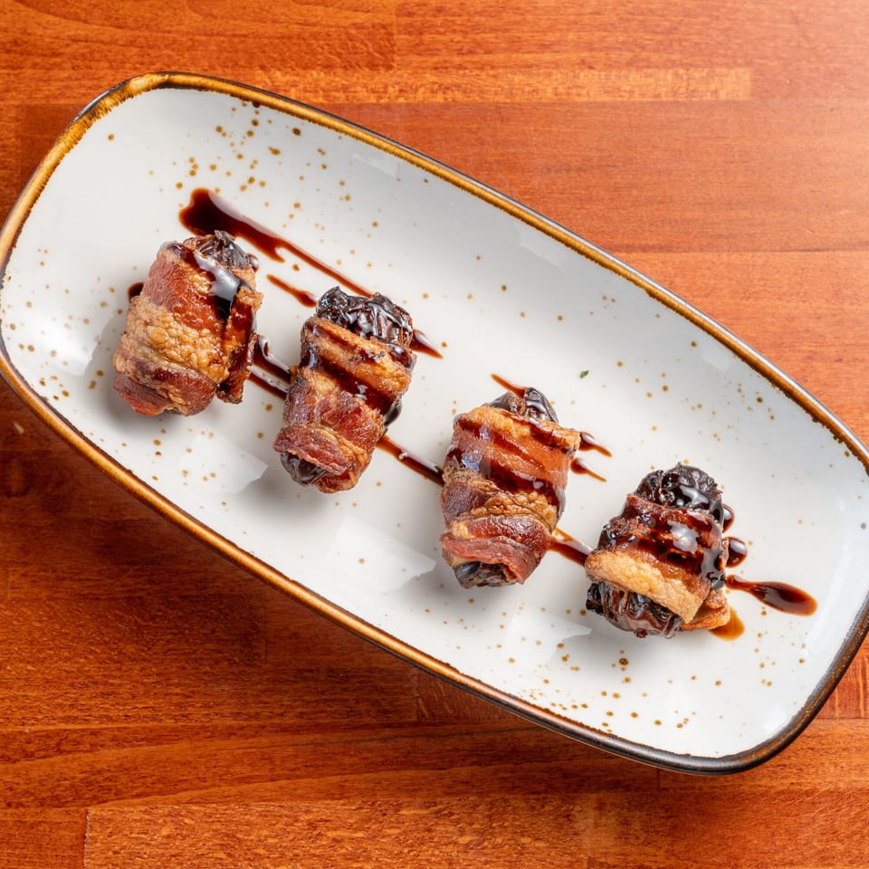Four bacon-wrapped dates drizzled with balsamic glaze on a white rectangular plate.