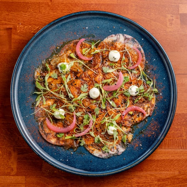 Taberna Ole's Spanish dish with sliced meat, red onions, microgreens, and white dollops on a blue plate.